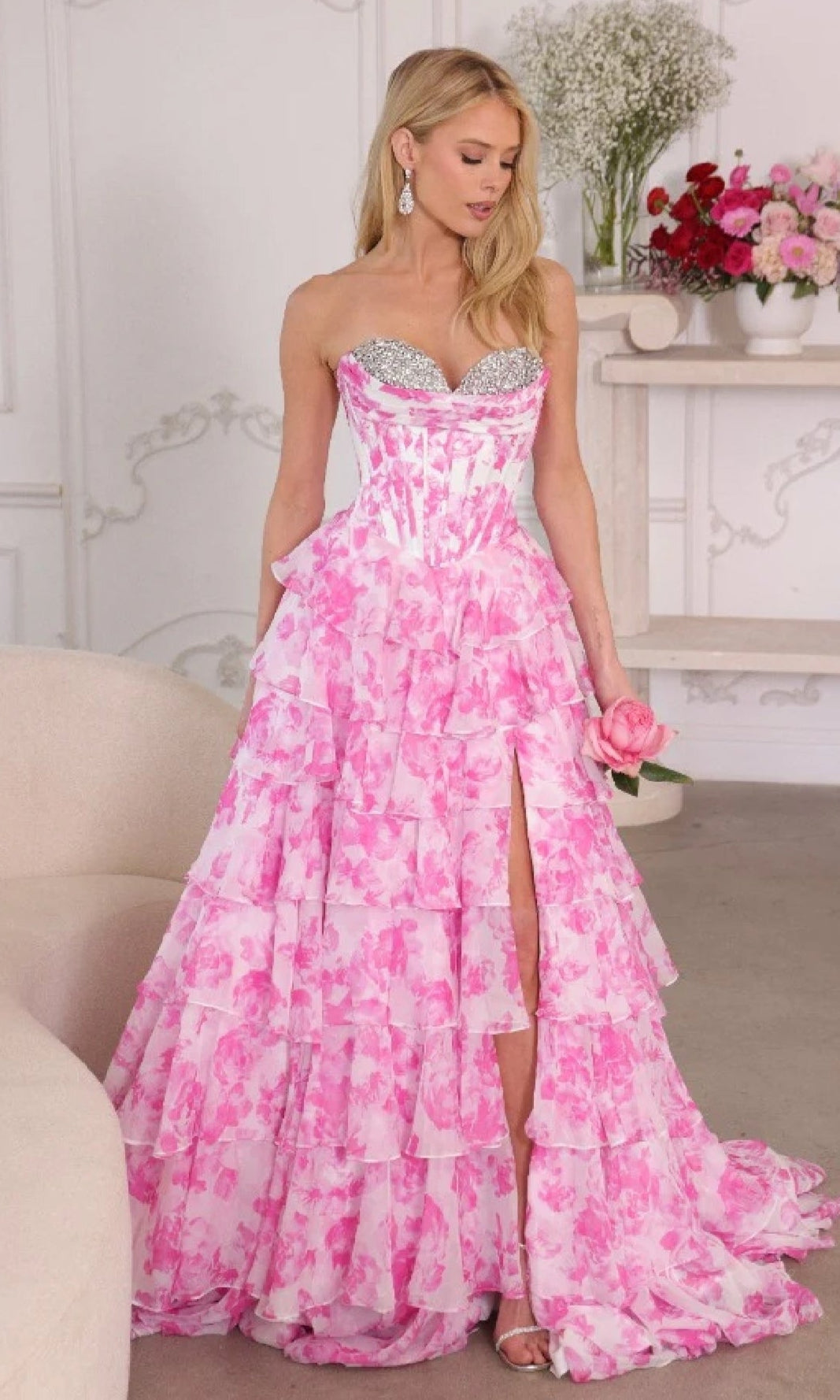 A woman wearing the Ava Presley 26441 long prom dress, with a strapless sweetheart corset bodice, pink and white floral ruffle ball-gown skirt, and thigh-high slit, holds a pink rose indoors.